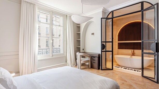 Une chambre moderne dotée d’un grand lit, d’un bureau et d’une salle de bain fermée par des parois de verre, avec une baignoire sur pieds.