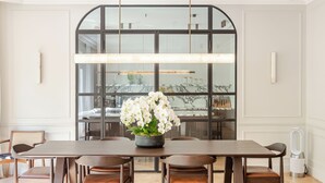 Un coin repas avec une longue table, six chaises, un vase de fleurs blanches et une grande fenêtre donnant sur la cuisine.