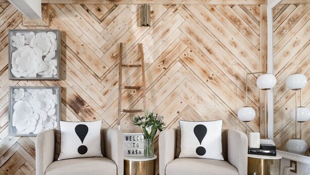 Un salon moderne avec un mur en bois à motifs en chevrons, deux canapés ornés de coussins noirs et blancs, une échelle, un panneau d’accueil et un lampadaire.