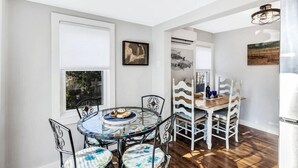 A dining area with a round glass table, metal chairs, and a wooden dining table with chairs. There is a window with blinds, a wall-mounted artwork, and a ceiling light fixture. - Falmouth Charm: Close to Main Street & Bike Trail RPAM098 (Falmouth)