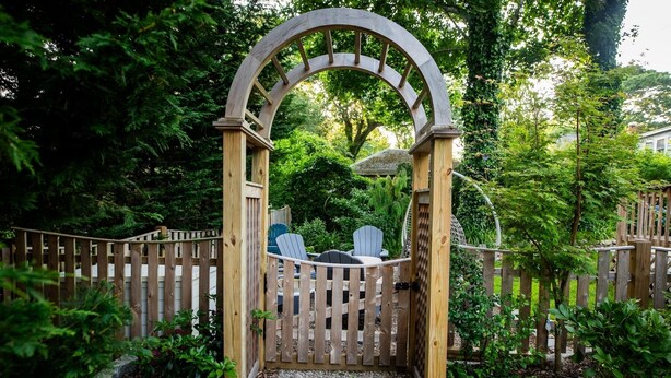 A wooden archway leading to a seating area with Adirondack chairs, surrounded by a wooden fence and greenery. - Falmouth Charm: Close to Main Street & Bike Trail RPAM098 (Falmouth)