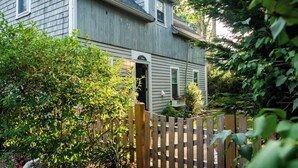 A two-story house with a wooden fence, surrounded by lush greenery and a small garden. - Falmouth Charm: Close to Main Street & Bike Trail RPAM098 (Falmouth)