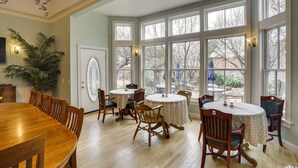 A dining area with a large wooden table and chairs, surrounded by large windows with white frames.