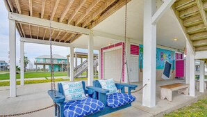 A covered porch with two blue swing seats labeled "Beach Blanket Bliss," a wooden bench, and a mural on the wall. - Steps to Beach: Pet-friendly Crystal Beach Home (Port Bolivar)