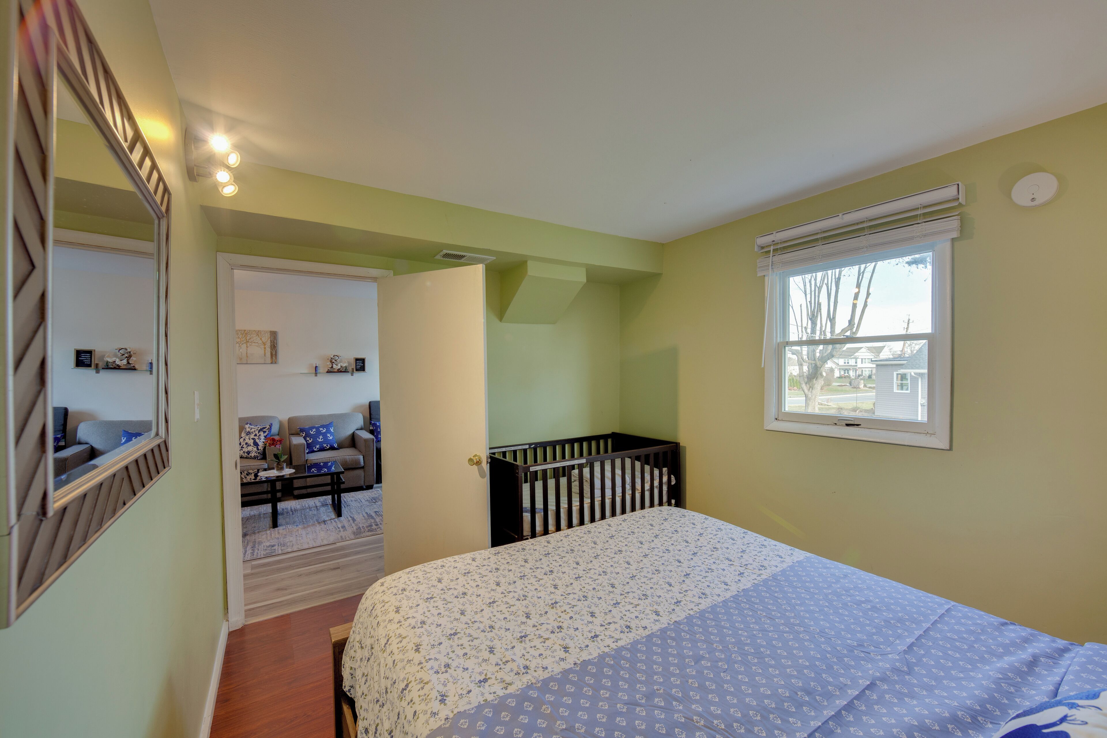 A bedroom with a bed, a window, a crib, a door leading to another room, and a mirror.