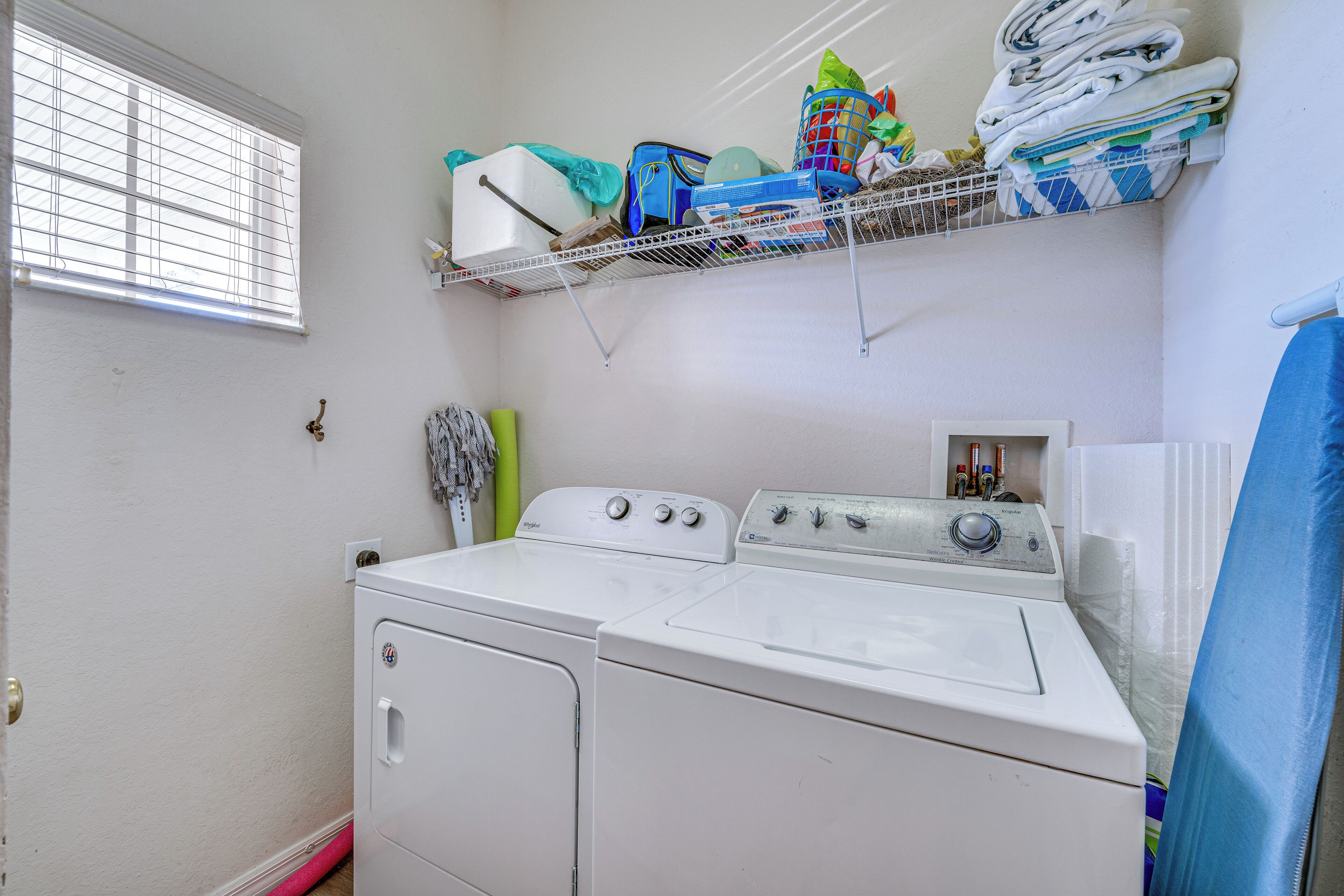 A laundry room with a front-loading washer and dryer, a shelf with folded towels and laundry baskets, and a window with blinds.