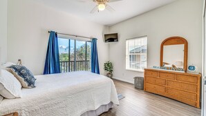 Ein Schlafzimmer mit Bett, Kommode, Deckenventilator, Fenster mit Vorhängen und Blick ins Freie.