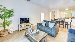 Ein geräumiges Wohnzimmer mit grauem Sofa, einem Holztisch und einem Fernseher. Ein Essbereich mit Tisch und Stühlen sowie eine Küche im Hintergrund sind ebenfalls zu sehen.
