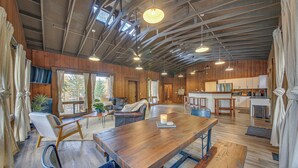 A spacious living area with a wooden dining table, comfortable seating, a kitchen area, and large windows providing natural light.