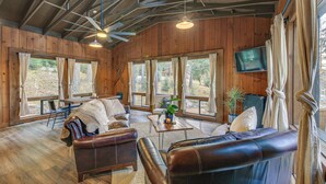 A spacious living room with leather sofas, a coffee table, and a TV mounted on the wall. Large windows allow natural light to fill the room.
