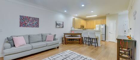 A modern living room with a grey sofa, a coffee table, and a dining area with a wooden table and chairs. The kitchen is visible in the background with wooden cabinets and a refrigerator.
