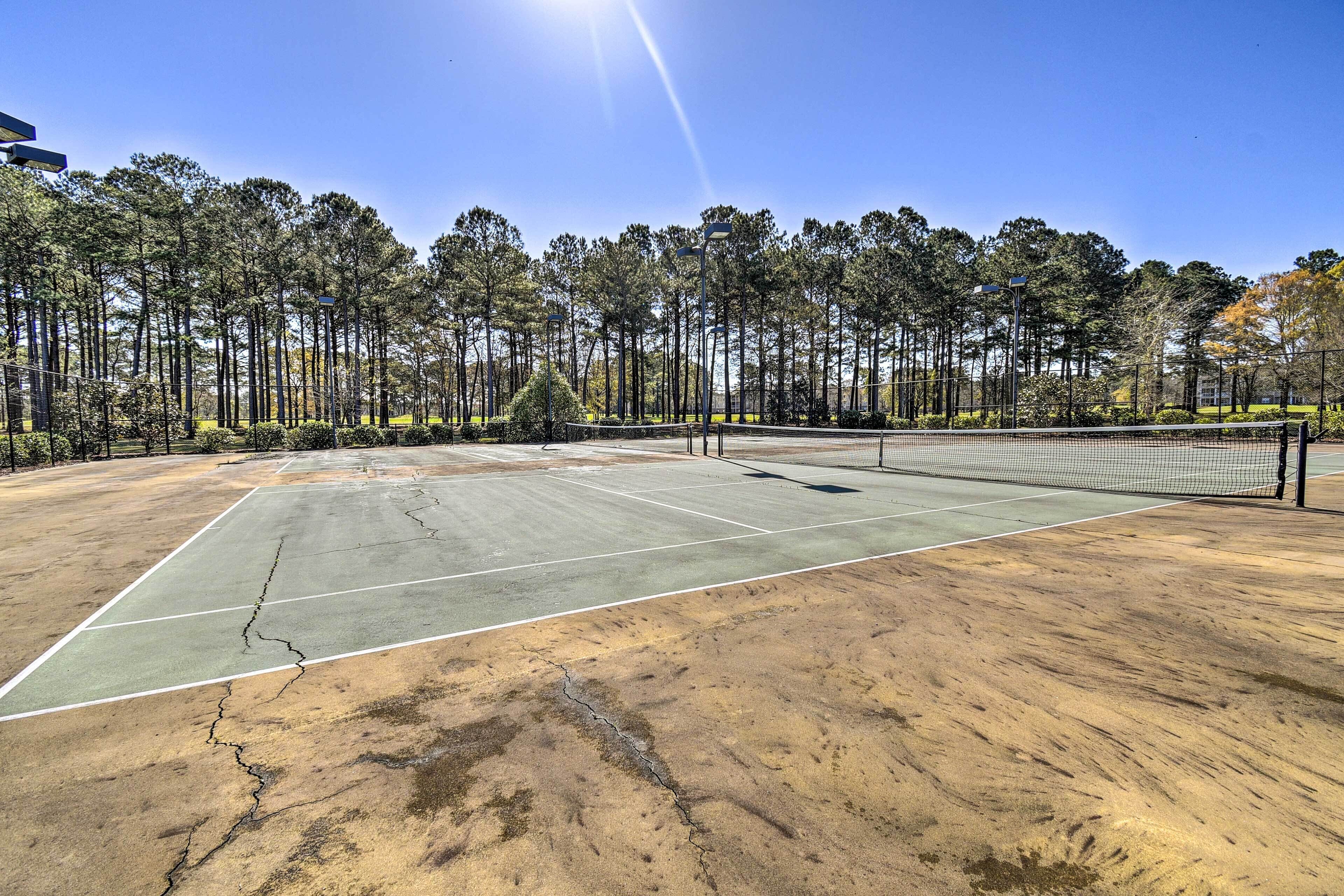 Una cancha de tenis con grietas visibles y una red, rodeada de árboles y un cielo despejado.