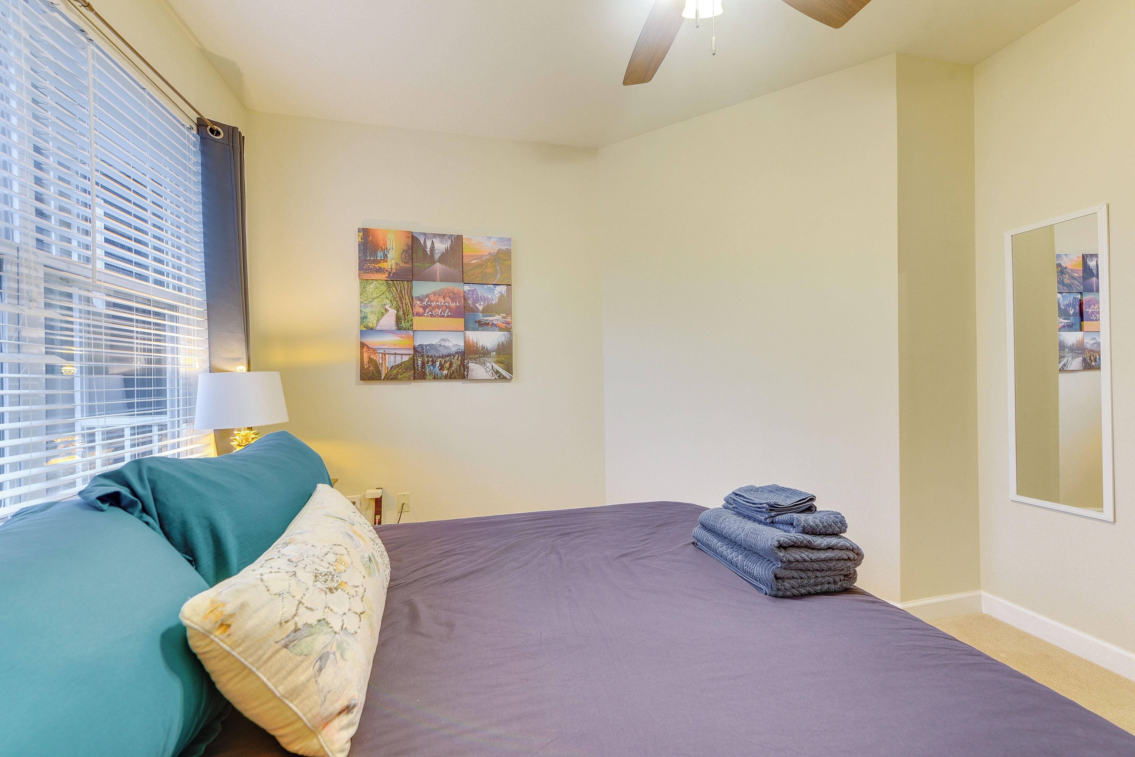 A bedroom with a bed, a window with blinds, a wall-mounted artwork, and a stack of towels.