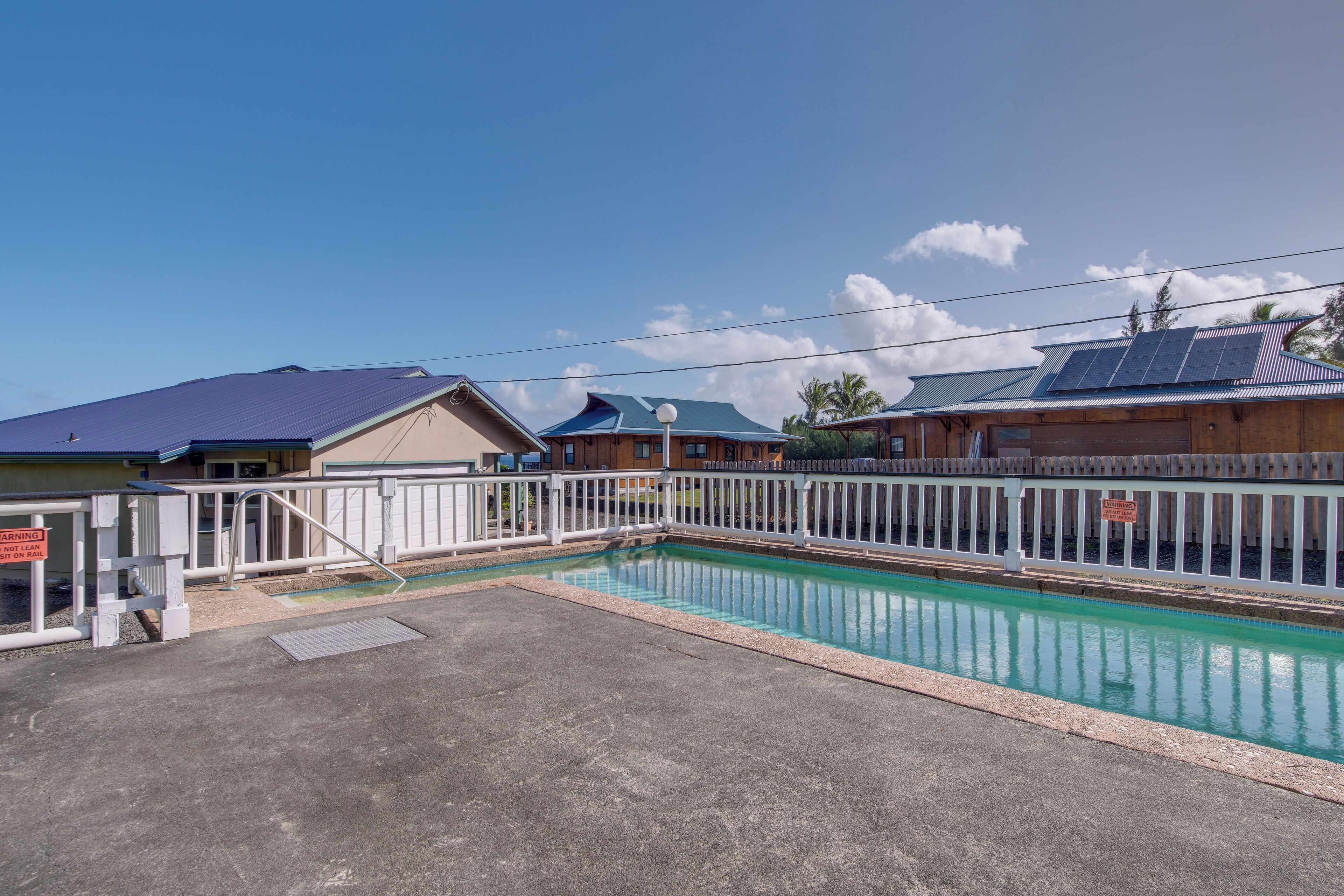 A swimming pool with a white fence, a covered area, and buildings with solar panels in the background.