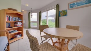 A dining area with a round table and wicker chairs, a bookshelf with books, and a view of greenery through large windows. - Oceanfront Keaau Gem: Ocean & Sunrise Views, Pool (Keaau)