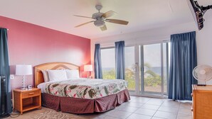 A bedroom with a bed, a ceiling fan, a nightstand, a lamp, a dresser, and a view of the outdoors through a sliding glass door. - Oceanfront Keaau Gem: Ocean & Sunrise Views, Pool (Keaau)