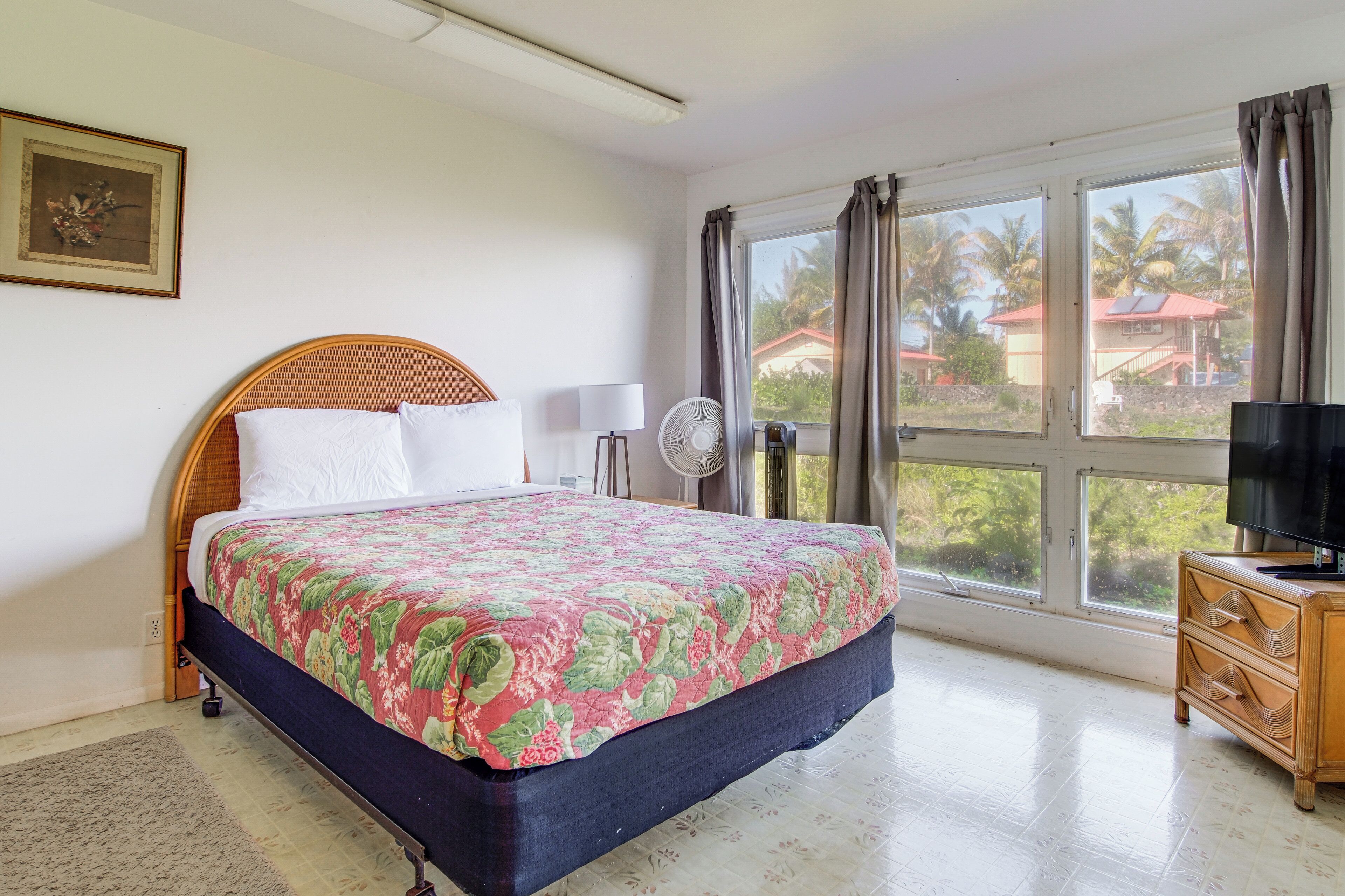 A bedroom with a bed, a bedside table with a TV, a window with curtains, and a wall-mounted fan.