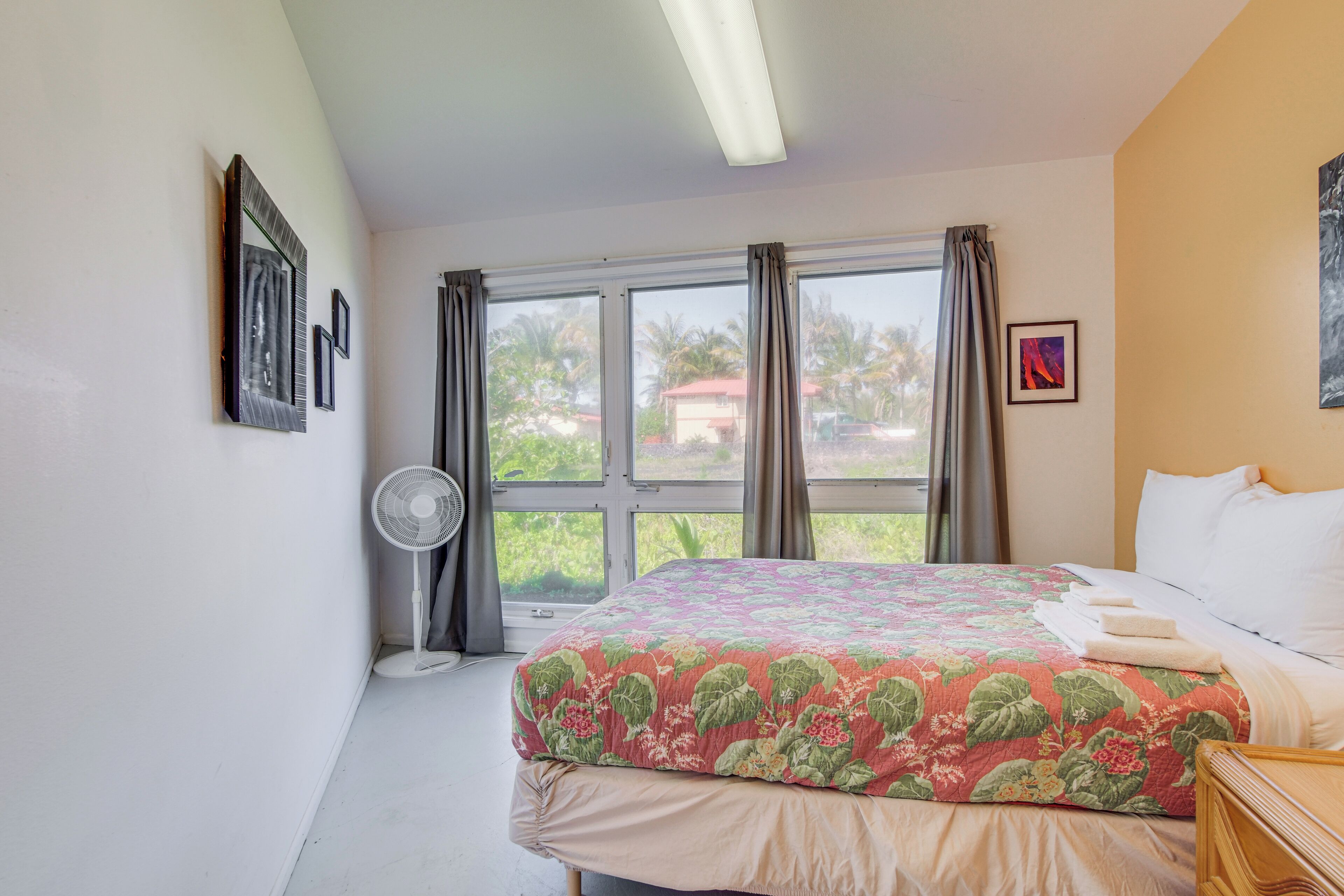 Ein Schlafzimmer mit einem Bett, einem Standventilator, einem Fenster mit Vorhängen und Blick ins Grüne.