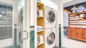 A laundry area with stacked washing machines, shelves with towels and laundry supplies, and a wooden cabinet with dishes. - Pet-friendly Garden Studio ~ 25 Mi to Boston! (Scituate)