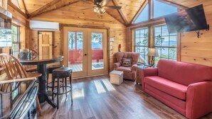 A wooden cabin interior with a dining table, chairs, a red sofa, a television, and a ceiling fan. - Majestic Views-Hot Tub - Views - Gas Firepit -4WD-AWD (Whittier)