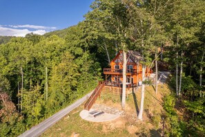 A two-story cabin with a wooden deck and a stone foundation, surrounded by dense forest. - Majestic Views-Hot Tub - Views - Gas Firepit -4WD-AWD (Whittier)