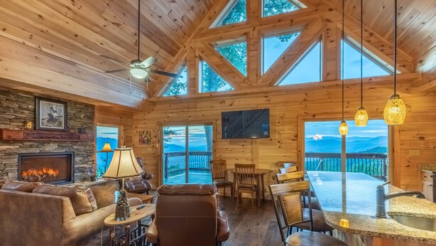 A cozy cabin interior with a stone fireplace, leather furniture, a dining area, and large windows offering mountain views. - Majestic Views-Hot Tub - Views - Gas Firepit -4WD-AWD (Whittier)