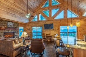 A cozy cabin interior with a stone fireplace, leather furniture, a dining area, and large windows offering mountain views. - Majestic Views-Hot Tub - Views - Gas Firepit -4WD-AWD (Whittier)