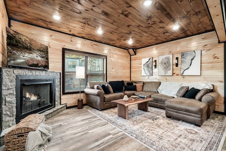 A cozy living room with a fireplace, wooden walls, and a large window.