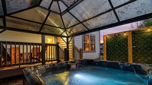 Un espace avec un bain à remous, entouré de parois en verre, doté d’une terrasse en bois et offrant une vue sur une maison et la verdure environnante.