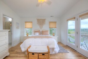 A bedroom with a bed, bedside tables, a ceiling fan, a window with blinds, and a balcony view. - Discover Oak Island's "Marsh to the Beach" - a spacious 3BR, 3BA haven for 8 near coastal fun! (Oak Island)