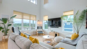 Ett rymligt vardagsrum med en stor soffa, ett soffbord och en tv som är monterad på väggen. Det finns stora fönster med persienner, en krukväxt och utsikt över omgivningen.