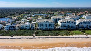 Un hotel frente a la playa con varias edificaciones, una playa de arena y vista a la zona residencial circundante.