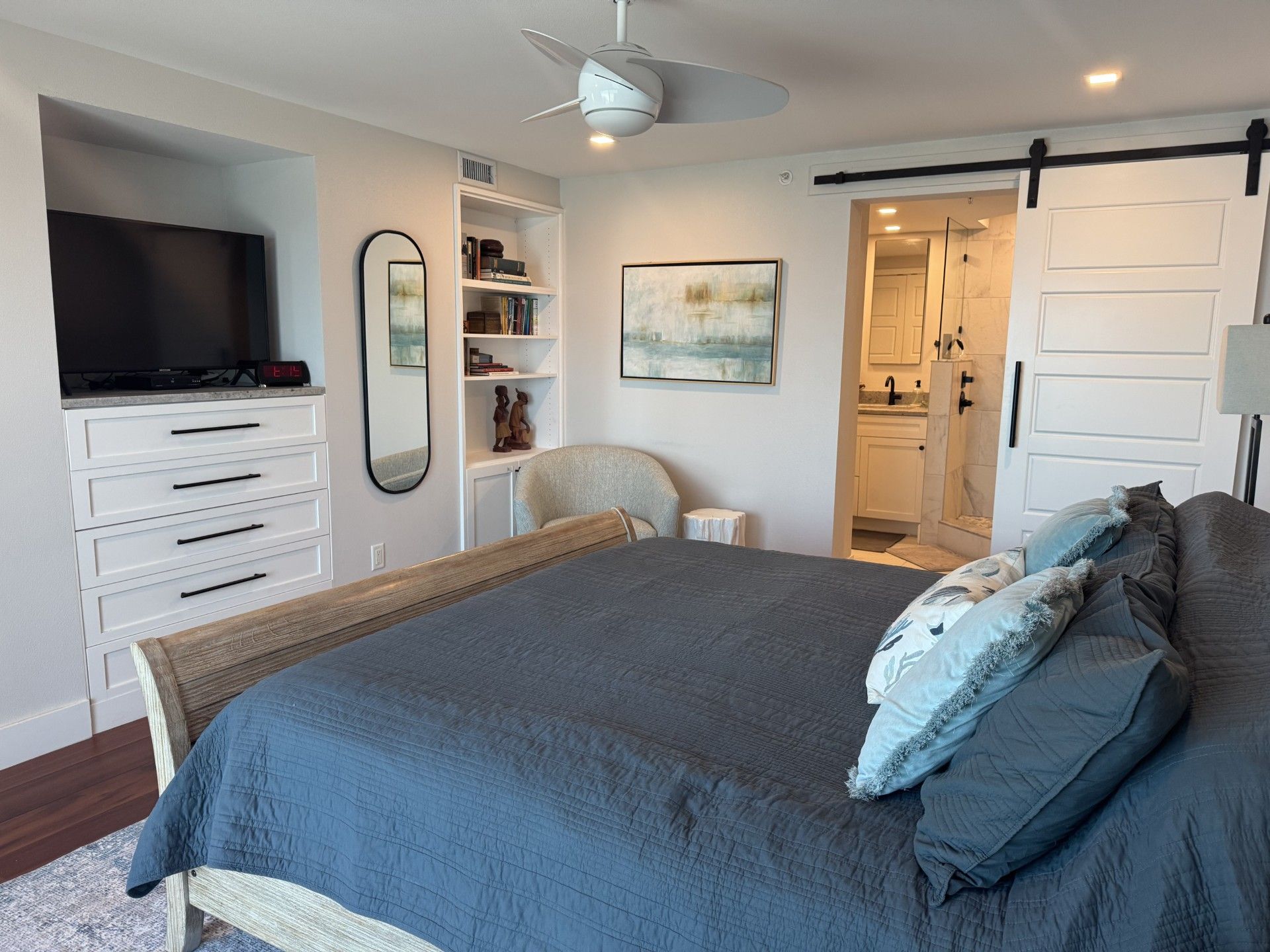 A bedroom with a bed, a television, a bookshelf, a chair, and a bathroom visible through an open door.