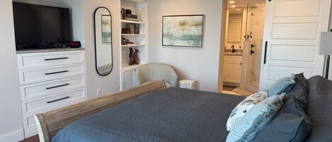 A bedroom with a bed, a television, a bookshelf, a chair, and a bathroom visible through an open door.