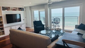 Ein Wohnzimmer mit Sofa, Couchtisch und Fernseher an der Wand. Ein Balkon bietet Blick auf den Ozean.