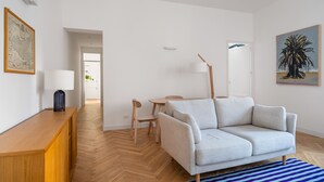 Un salon moderne avec un canapé gris clair, une table basse en bois et des fauteuils. On y trouve une table d’appoint en bois avec une lampe, un tapis à rayures bleues et des œuvres d’art accrochées aux murs.
