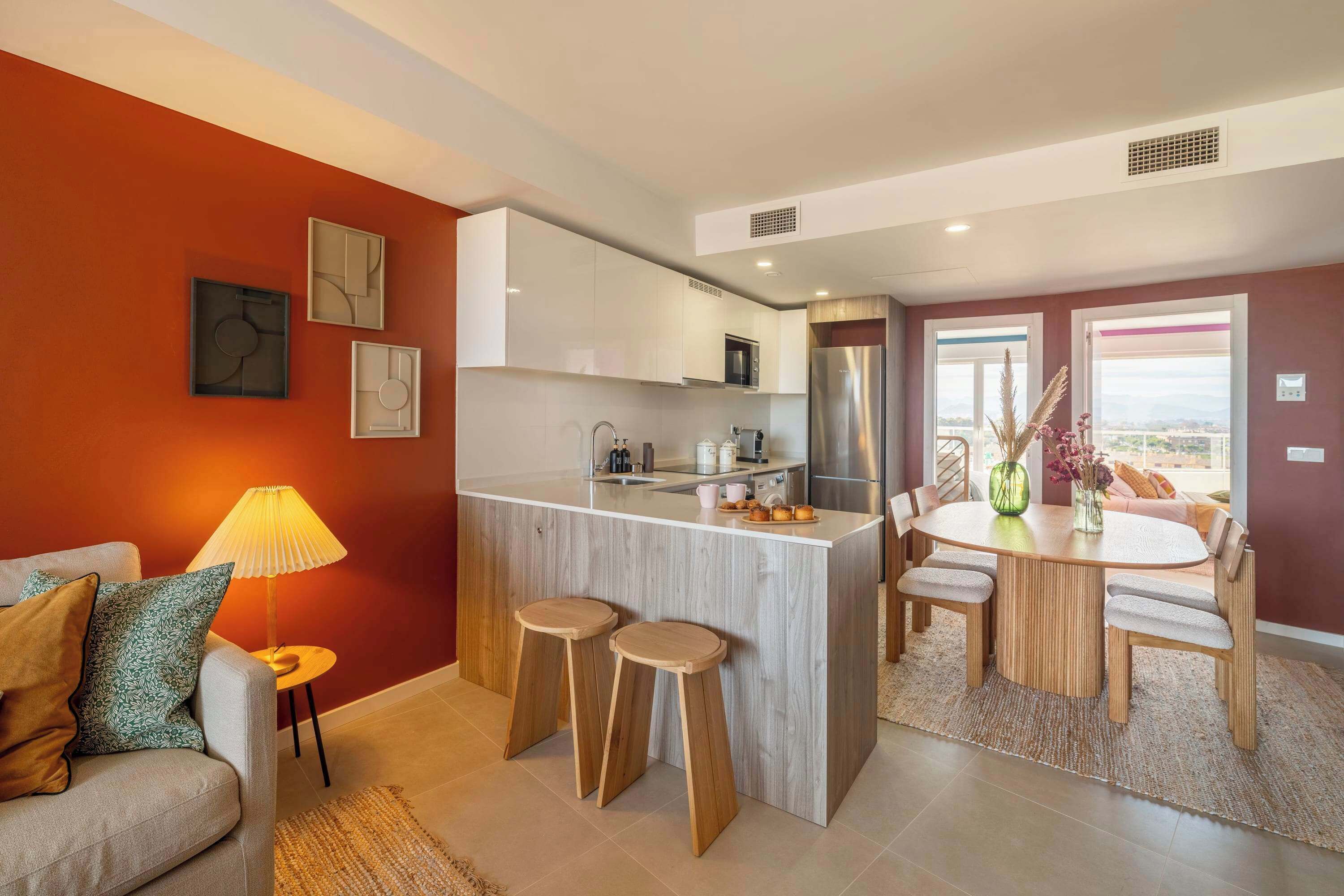 Una cocina moderna con armarios blancos, una isla central con taburetes, un comedor con una mesa redonda y sillas, y un sofá con lámpara.