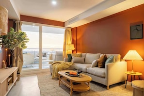 A modern living room with a sofa, coffee table, and a lamp. There is a large window with curtains, a plant, and a view of the outdoors.