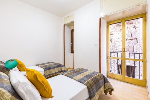 Una habitación con dos camas, una ventana que da a un muro de piedra y una puerta que conduce a otra habitación.