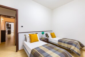 Hotellihuone, jossa on kaksi sänkyä, pieni työpöytä, lamppu ja näkymä keittiöön.