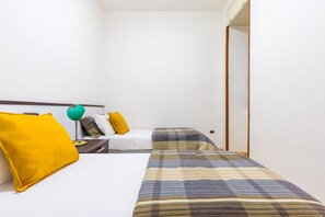 Habitación de hotel con dos camas, una con cubrecama a cuadros y la otra con cubrecama blanca lisa. Hay una mesita de noche con una lámpara y un cojín amarillo.
