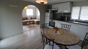 Et moderne køkken med hvide skabe, en mikrobølgeovn og en komfur. Der er et spiseområde med et rundt bord og stole, og en sofa i baggrunden.