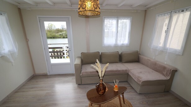 Et værelse med trægulv, en beige sofa, et lille bord med en vase og et stearinlys, samt udsigt gennem en dør til et udendørs område.