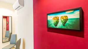 A room with a red wall, a mounted TV displaying a landscape, a seating area with chairs, and a wall-mounted air conditioning unit. - Central Station Apts by Wonderful Italy - Capri (Napoli)