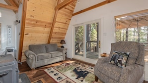 Ein gemütliches Wohnzimmer mit Holzboden, einem grauen Sofa, einem gemusterten Sessel und einem großen Fenster mit Jalousien.