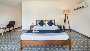 Una habitación de hotel con una cama de madera, una mesita de noche pequeña con una lámpara, una silla y baldosas de suelo estampadas.