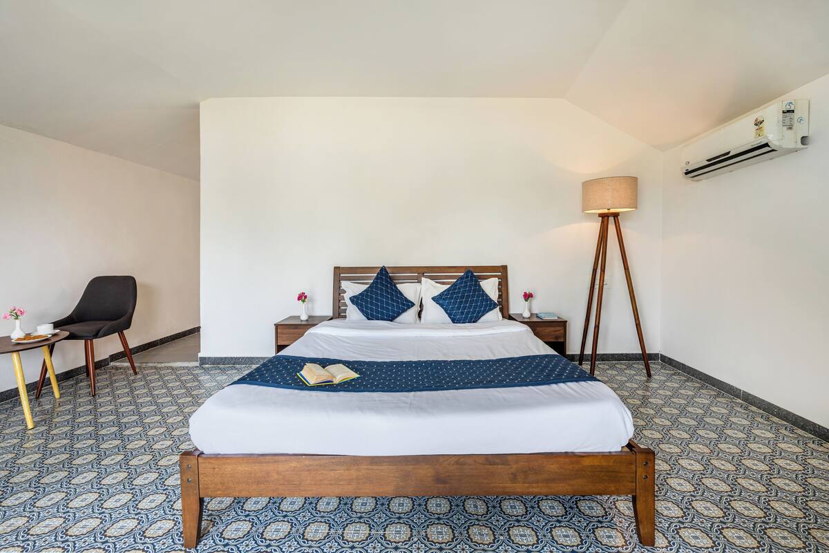 Una habitación de hotel con una cama de madera, una mesita de noche pequeña con una lámpara, una silla y baldosas de suelo estampadas.