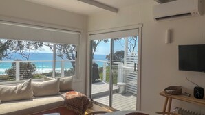 A modern living room with a large window overlooking the sea, a sofa, a table, and a TV mounted on the wall. - PIPI SHACK Iconic waterfront shack at Binalong Bay Beach, Bay of Fires (Binalong Bay)