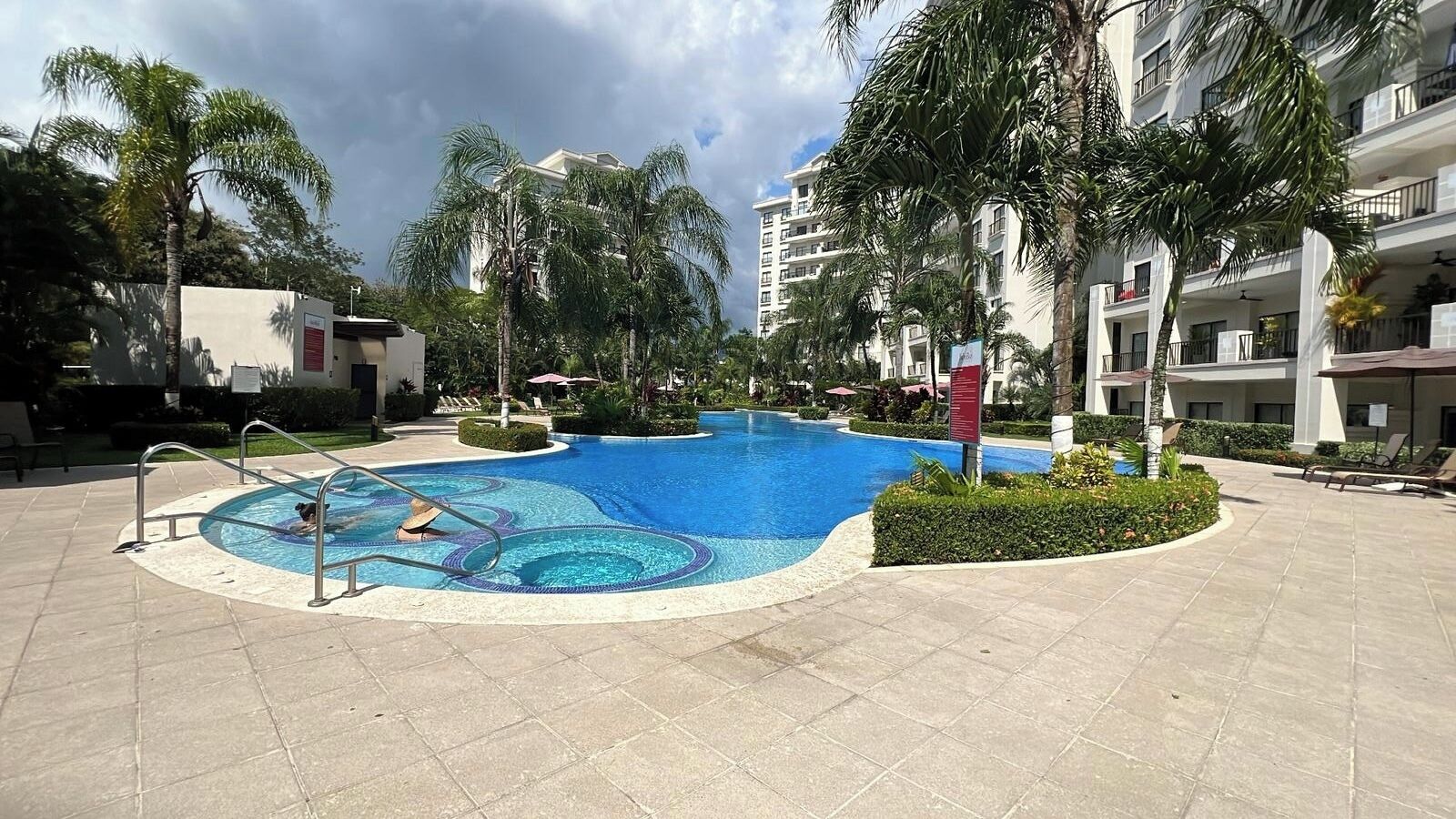 A swimming pool with a circular design, surrounded by palm trees and a building with balconies.