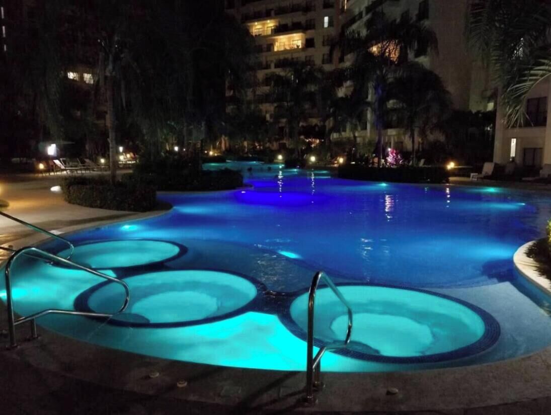 A nighttime swimming pool with circular patterns, surrounded by palm trees and hotel buildings.